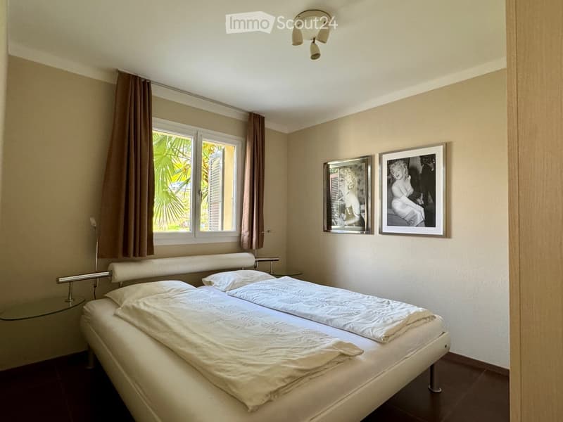 Modern bedroom with a double bed, nightstand, glass table, and two framed pictures on the wall. The room features large windows with curtains and a ceiling light fixture.