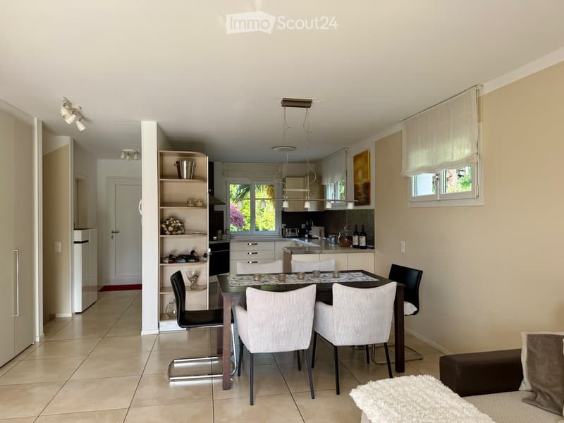 Modern dining area with a dark wooden dining table, six chairs, and a small open shelving unit with decorative items. The adjacent kitchen features white cabinets, stainless steel appliances, and a countertop with jars and bottles.