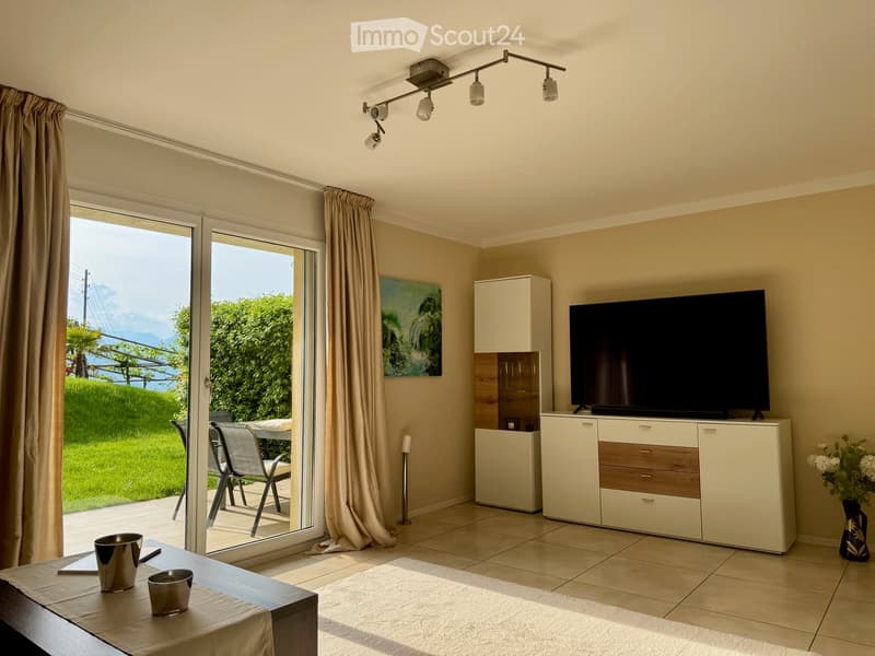 Bright living room with large sliding glass doors leading to a patio, a TV on a white cabinet, a small table with two silver cups, a painting on the wall, and a view of greenery and mountains.