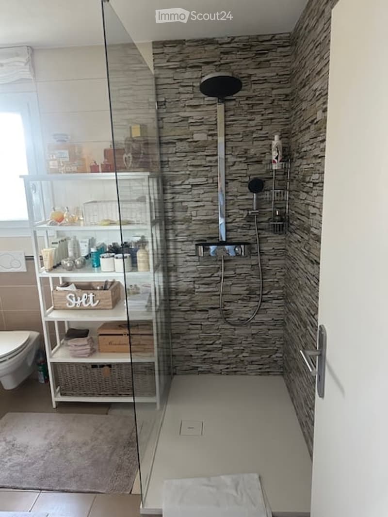 Modern bathroom with a glass-enclosed shower, stone-textured wall, and a white shelving unit holding toiletries and towels.