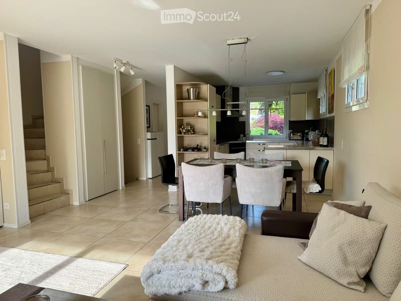 Modern living and dining area with a sofa, coffee table, dining table with chairs, and an open kitchen area with cabinets, appliances, and a window view.