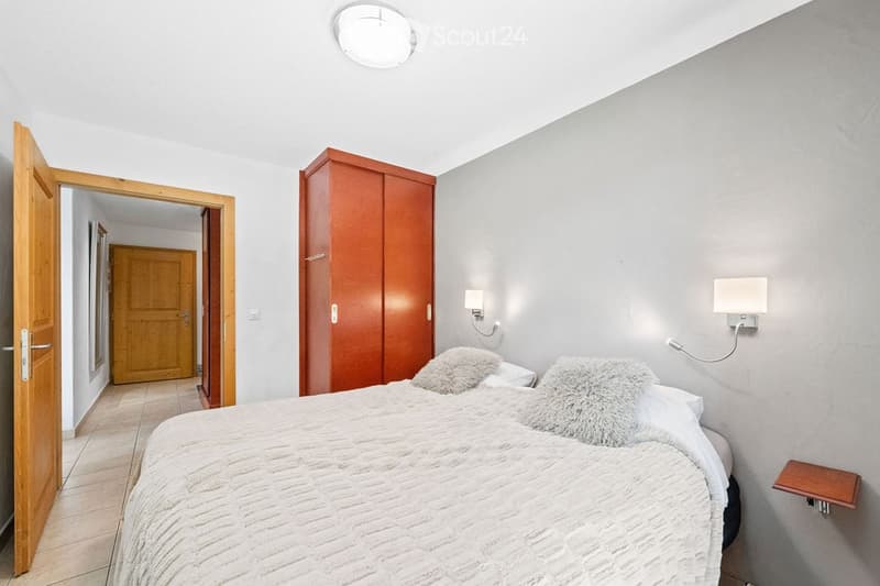 Modern bedroom with a large bed, fluffy pillows, a wooden wardrobe, and wall-mounted lamps. The room features light grey walls and a wooden door leading to a hallway.