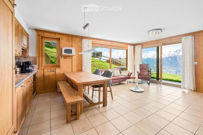 Open-plan living and dining area with wooden walls, large windows offering a mountain view, a red sofa, a wooden dining table with chairs, a bench, and a kitchen area with cabinets and appliances.