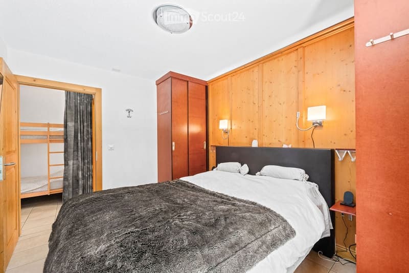 Cozy bedroom with a double bed, pillows, a fur throw, a wardrobe, a nightstand with a lamp, and a mirror. The room features wooden paneling and a bunk bed in the adjacent space.