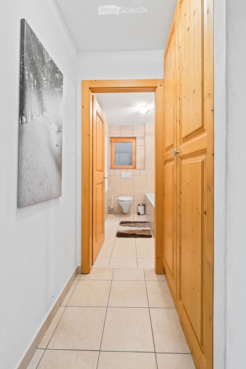 A hallway with tiled flooring leads to a small bathroom. The bathroom features a toilet, a small window, a rug, and a trash bin.