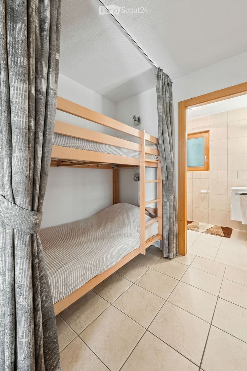 Cozy bedroom with a wooden bunk bed, neatly made with bedding, and a curtain on one side. A small adjacent bathroom is visible with a tiled floor and a window.