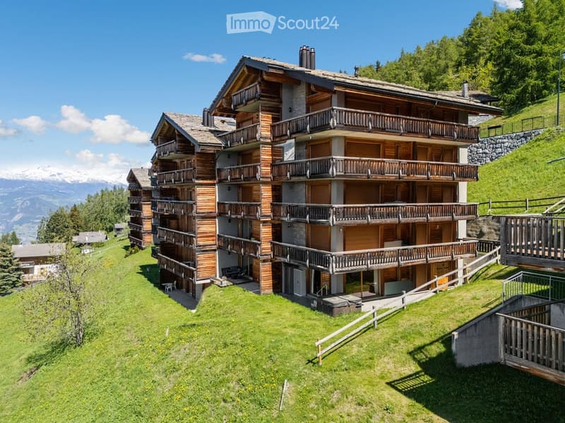 Multi-story wooden building with balconies, surrounded by green lawn and mountains in the background.