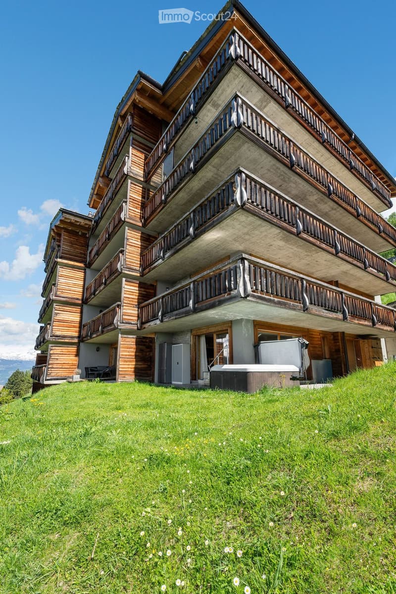 A multi-story wooden house with balconies, situated on a grassy slope with a view of mountains in the background.
