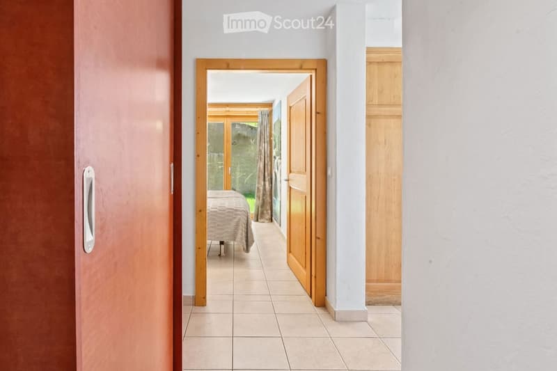 A hallway with tiled flooring, white walls, and wooden doors leading to a bedroom and a room with large windows and a view of greenery.