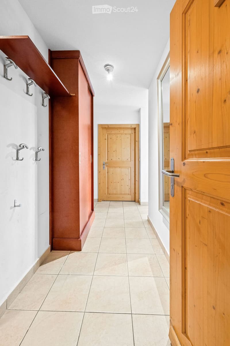 A clean, well-lit hallway with tiled flooring, white walls, and wooden doors. Features include built-in wooden shelving on one wall and coat hooks.