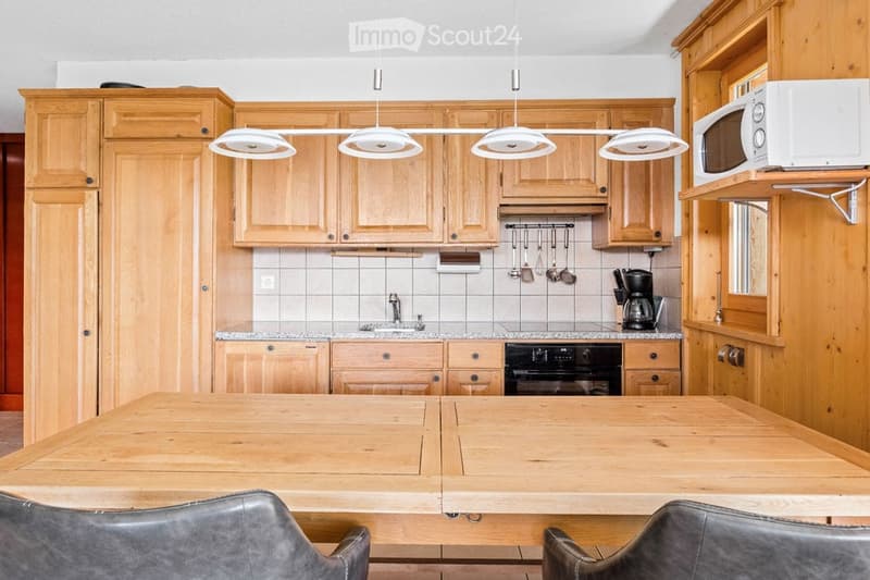 Bright kitchen with light wooden cabinets, granite countertops, and a central wooden dining table with leather chairs. Appliances include a microwave, coffee maker, and oven.