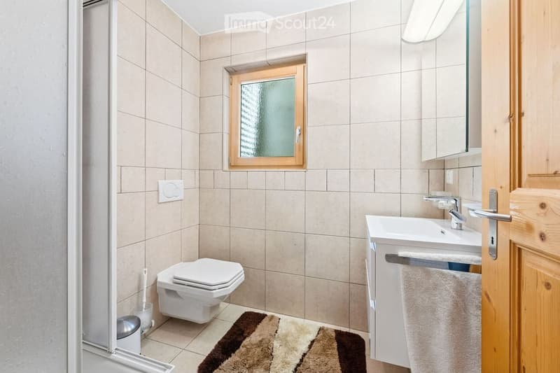 A modern bathroom with beige tiled walls and floor, a toilet, a sink with a mirror, a towel, and a window with wooden frame.