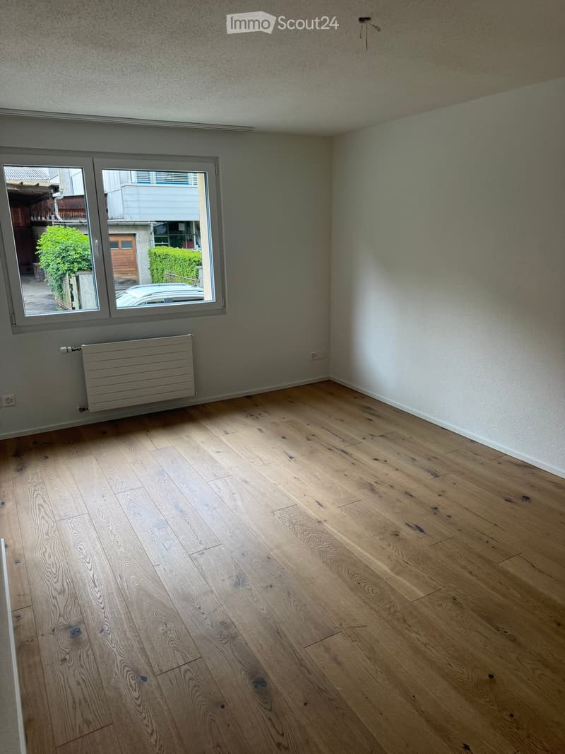 Empty room with wooden flooring, white walls, and a large window with a view of the outside. A radiator is visible below the window.
