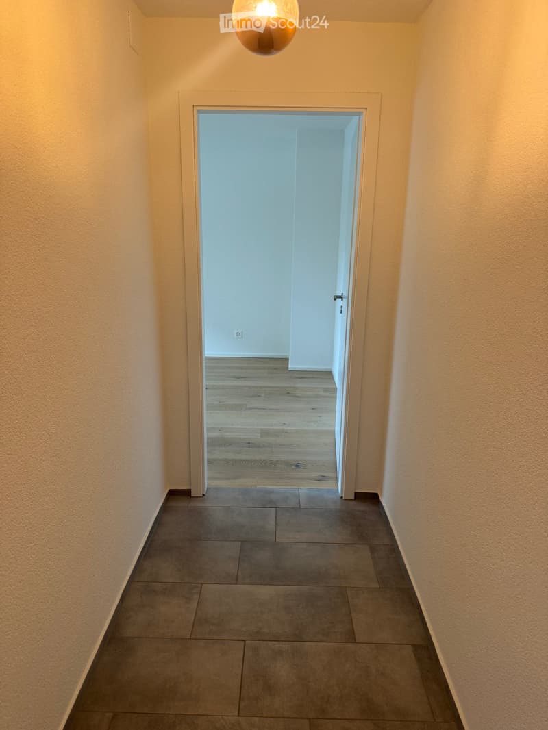 Empty hallway with beige walls, tiled floor, and a wooden floor visible in the adjacent room.