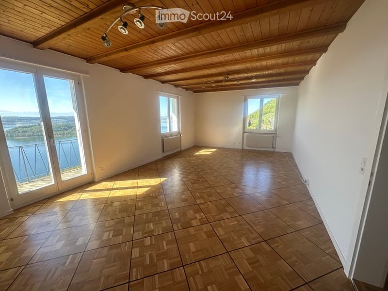 Empty room with wooden herringbone flooring, white walls, large windows offering scenic views, and exposed wooden ceiling beams.