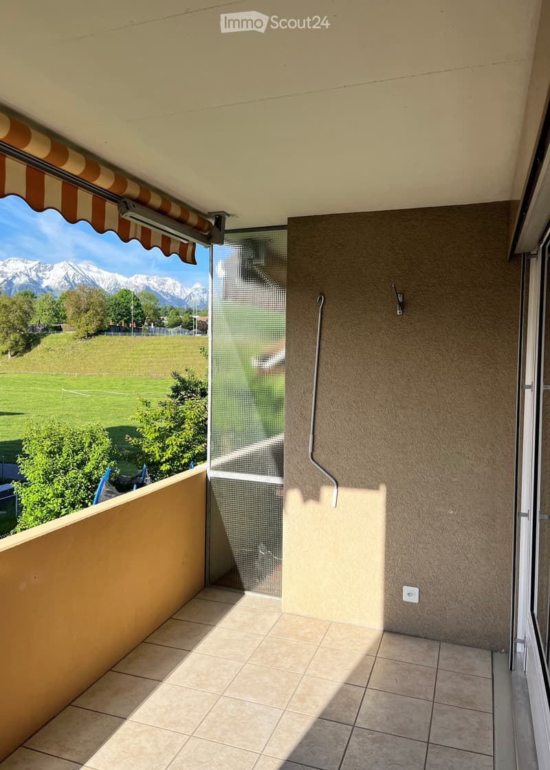 Balcony with tiled floor, beige walls, and a retractable awning, offering a scenic view of mountains and a green field.