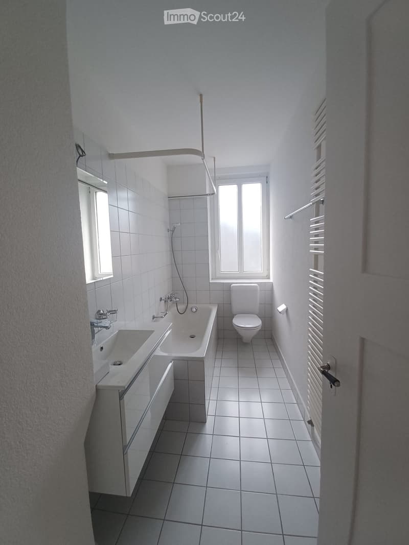 Modern bathroom with white tiles, a sink, bathtub, toilet, and radiator. Features a window with blinds and a glass shower enclosure.