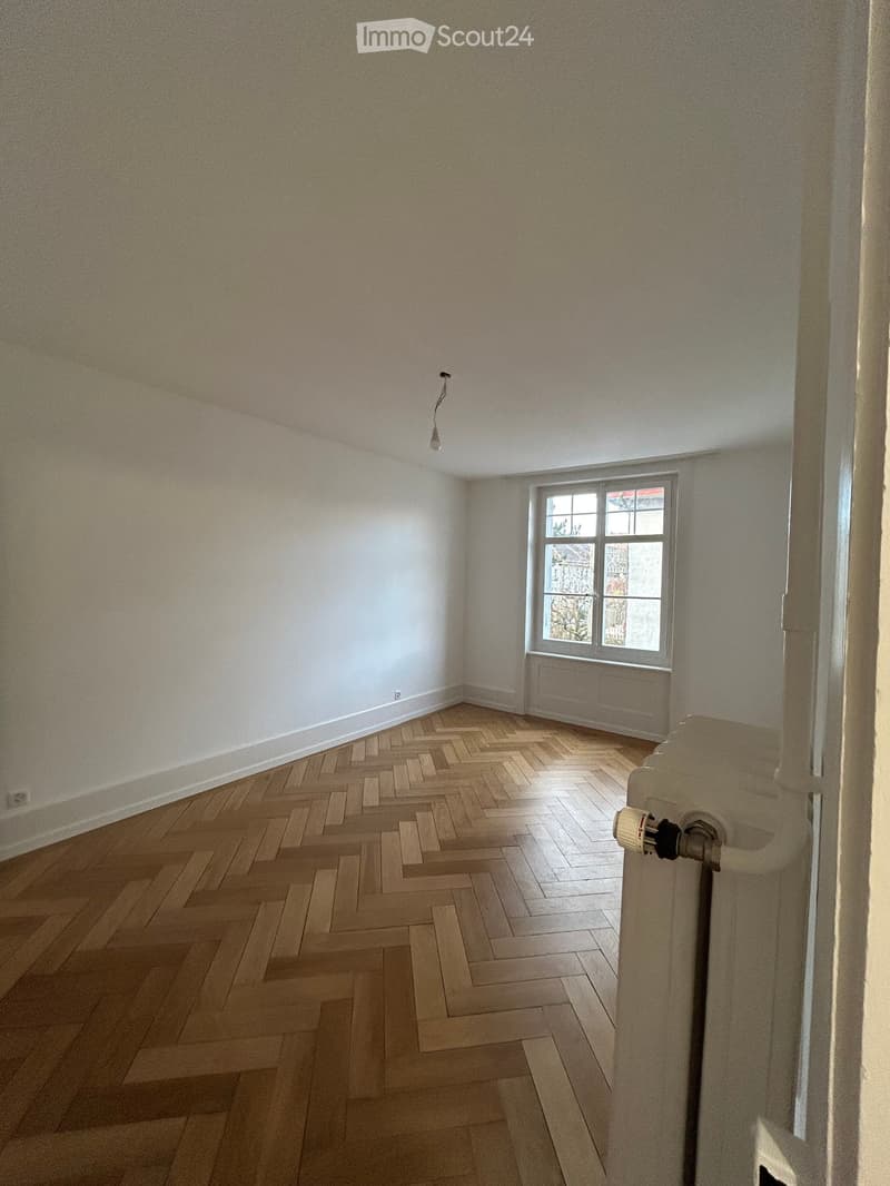 Empty room with wooden herringbone flooring, white walls, and a single window with a radiator below it. The room has a simple ceiling light fixture and a door frame visible on the right side.