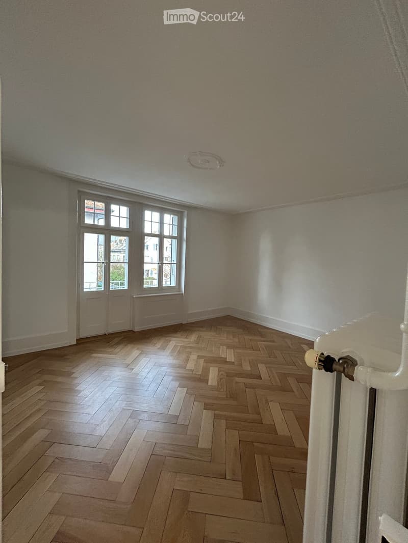 Empty room with wooden herringbone flooring, large windows, and a radiator on the wall.