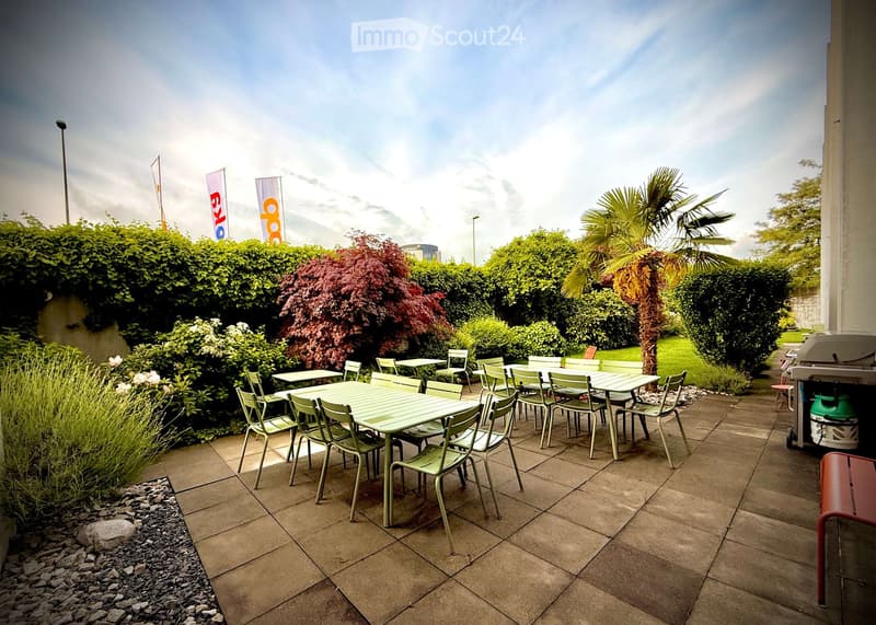 Outdoor seating area with tables and chairs arranged on a tiled patio, surrounded by greenery and bushes.