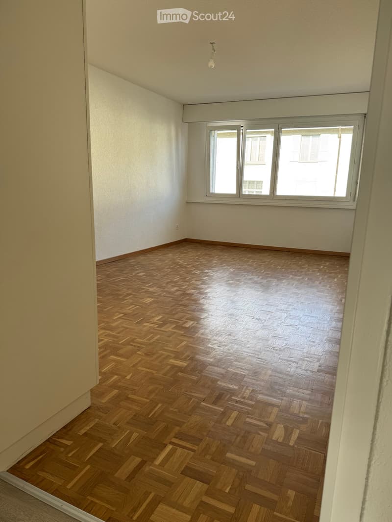 Empty room with wooden herringbone flooring, white walls, and large windows allowing natural light.
