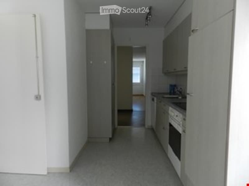 Empty hallway with white walls, grey carpeted floor, and a view into a kitchen area with cabinets and appliances.