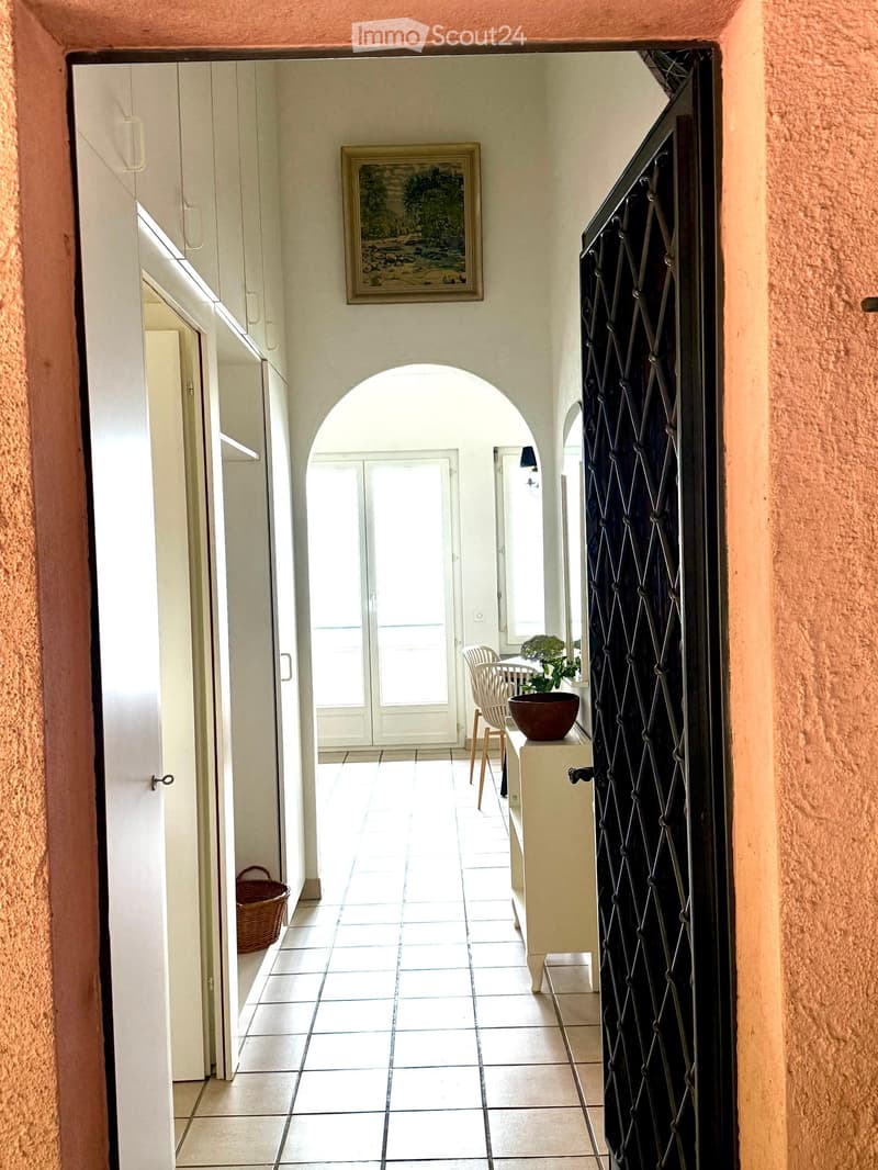 A well-lit hallway with tiled flooring, white walls, and an arched doorway leading to another room. A small table with a plant and a chair is visible in the adjacent room.