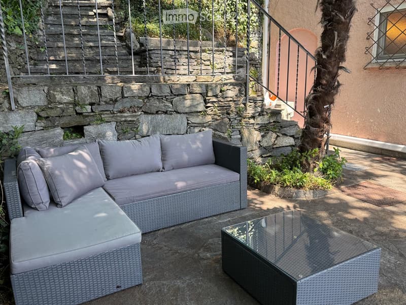 Outdoor seating area with a grey wicker sofa and matching coffee table, surrounded by stone walls and greenery.