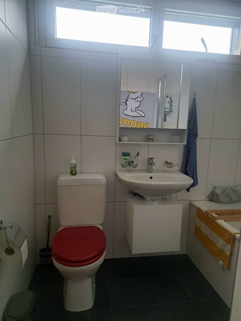 Bathroom with a toilet, sink, mirror, and various toiletries. The sink has a faucet and a toothbrush holder, and a towel hangs on the side. A bottle is placed on the toilet tank.