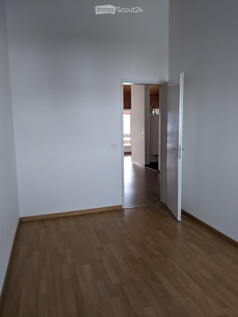 Empty room with light-colored walls and wooden flooring, featuring an open doorway leading to another room with visible furniture.