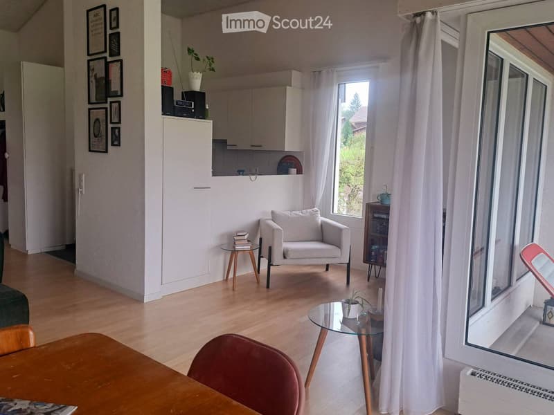 Bright living room with wooden flooring, white walls, and large windows. Features a grey armchair, glass coffee table, small wooden side table, and a dining table with chairs.