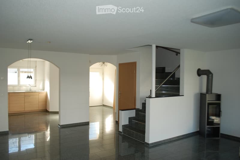 Bright, open hallway with polished tile flooring, white walls, and a staircase leading upwards. Features a wooden door, a small kitchen area with cabinets and sink, and a modern wood-burning stove on the right side.