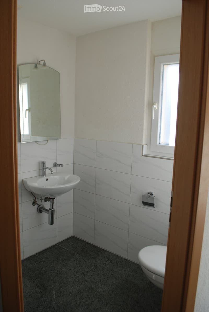 Modern bathroom with white tiles, a white sink, a chrome faucet, a mirror, and a toilet. The walls are painted in a light color, and the floor is covered with dark gray tiles.