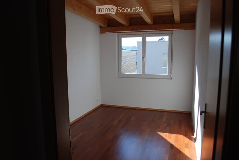 Empty room with wooden flooring and white walls, featuring a large window with blinds and a visible electrical outlet on the wall.