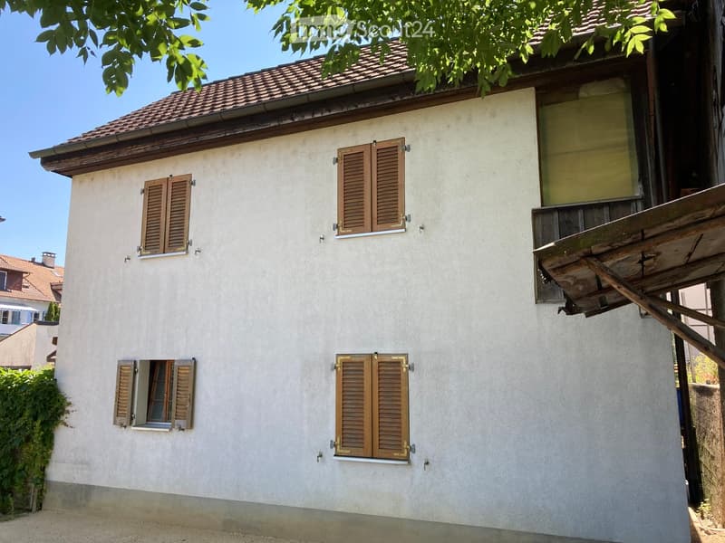 Two-story house with beige walls, wooden shutters, and a tiled roof. Features a wooden balcony on the right side.