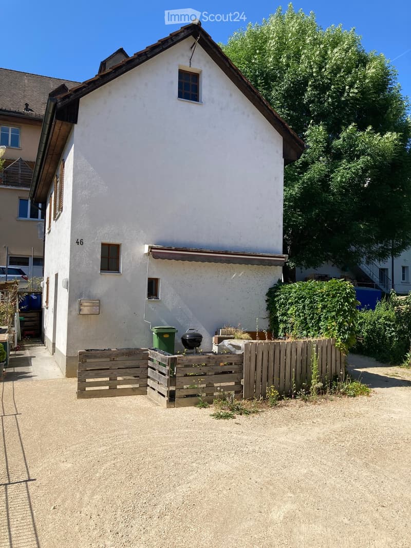 A two-story house with a white exterior and a brown roof, featuring a small garden area with a wooden fence, barbecue, and greenery.