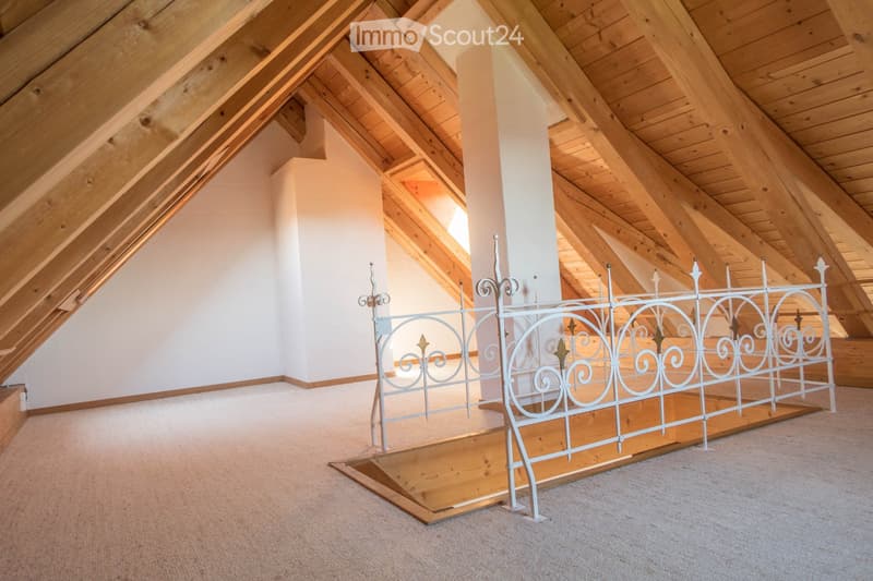 An empty attic space with wooden beams, white walls, and a metal staircase leading up, featuring exposed wooden beams and a carpeted floor.