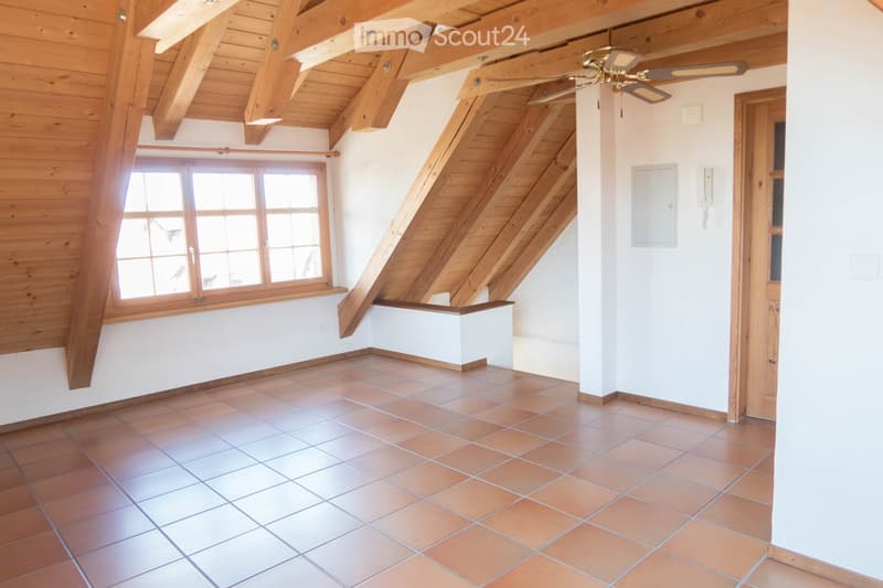 Empty room with exposed wooden beams, tiled floor, large window, and staircase leading upwards.