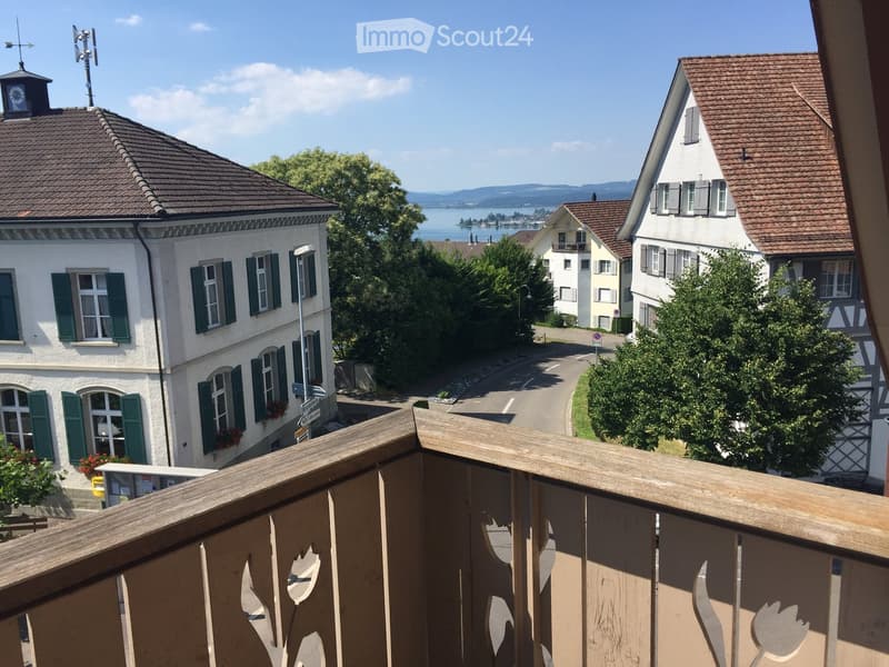 Balcony overlooking a street lined with traditional houses, with a scenic view of a lake and distant mountains.