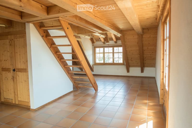 Empty room with wooden beams on the ceiling, tiled floor, and a wooden staircase leading upwards. The walls are white, and there is a window letting in natural light.