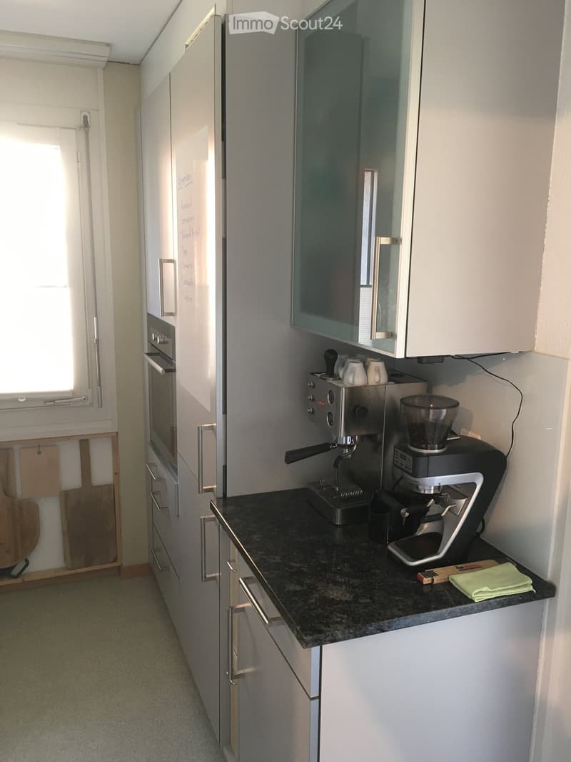 Modern kitchen with white cabinets, granite countertop, stainless steel appliances including a refrigerator, oven, and coffee machine. A glass-fronted cabinet is visible above the counter.