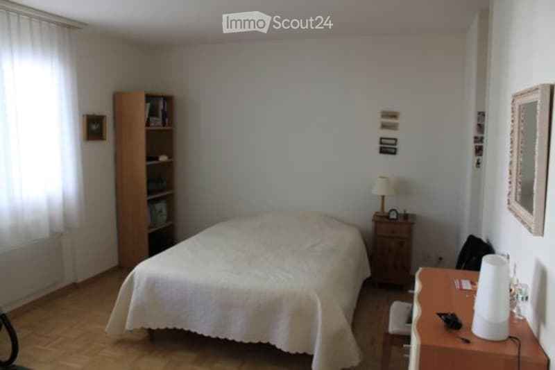 A tidy bedroom with a single bed, nightstand, desk, and bookshelf. The room features wooden flooring and white walls.