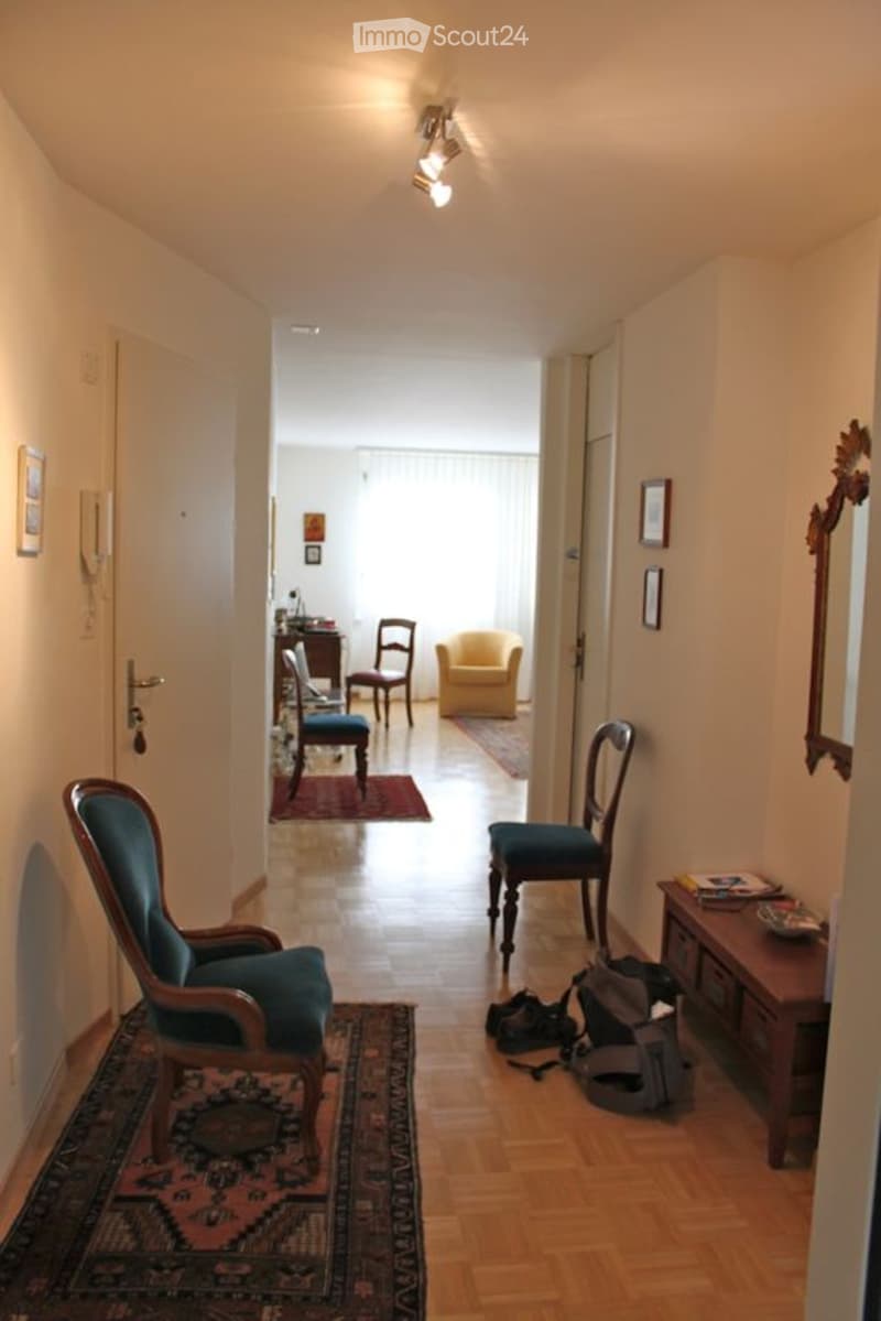 A well-lit hallway with wooden flooring, leading to a room with a yellow armchair, wooden chairs, and a small table. The hallway features a patterned rug and a small wooden side table with books and bags.