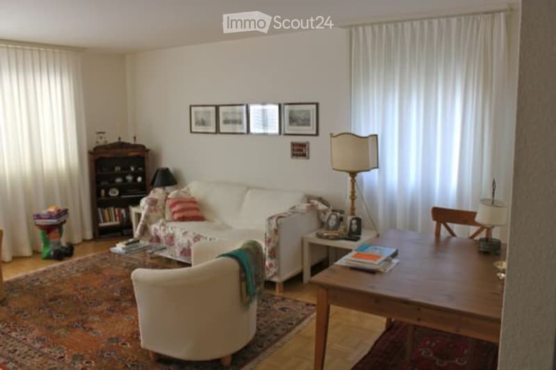 Bright living room with large windows covered by white curtains, a white sofa with colorful pillows, a wooden coffee table, a floor lamp, a bookshelf, and a small armchair.