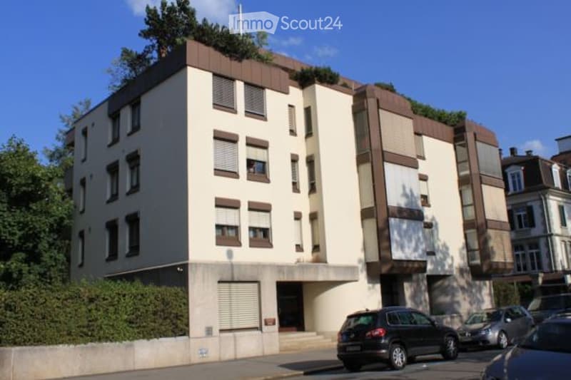 Modern multi-story building with a flat roof, white facade, and brown accents. Windows have shutters, and there is a small garden on the roof.