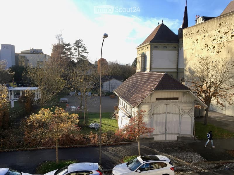 A view of a campus area with a large building featuring a tower and spire, surrounded by trees, a small shed, and parked cars.