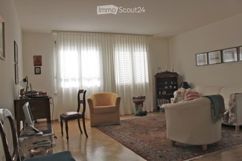 Bright living room with large windows covered by white curtains, a cream-colored sofa, a yellow armchair, a wooden side table, a bookshelf, and a computer desk with a monitor.
