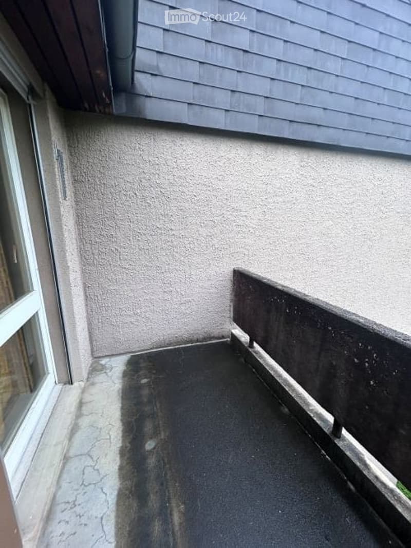 Small, enclosed balcony with a concrete floor, low concrete railing, and a window opening to the left.