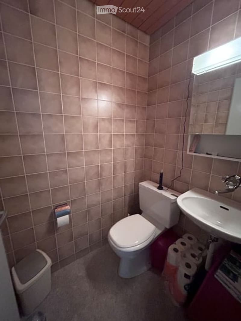 Small bathroom with beige tiled walls, a toilet, a sink with a mirror, and a trash bin. Toilet paper is placed near the toilet.