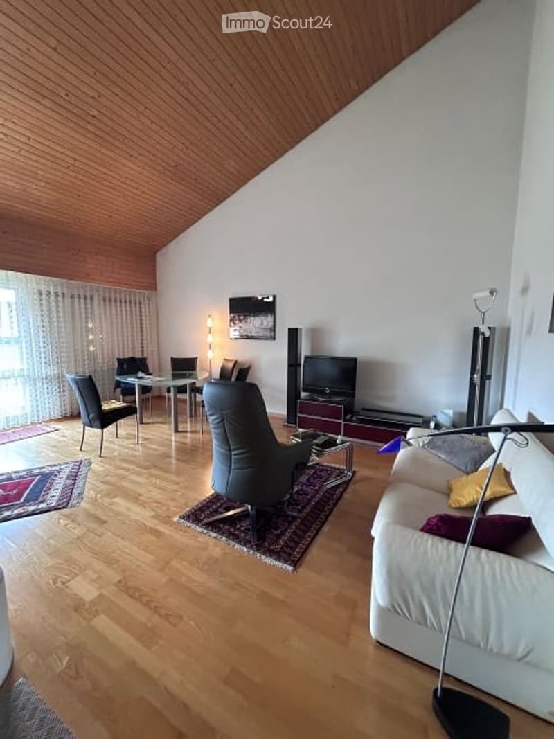 Modern living room with wooden flooring, white walls, and a slanted wooden ceiling. Features a white sofa with colorful cushions, a black leather armchair, a coffee table, a TV, and a dining area with chairs and a table.